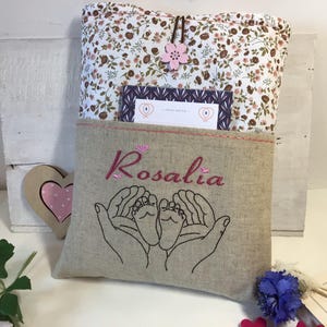 May include: A beige linen book cover with a floral pattern and a pink flower. The cover has a pocket with the name "Rosalia" embroidered in pink thread and an embroidered design of two hands holding a pair of baby feet.