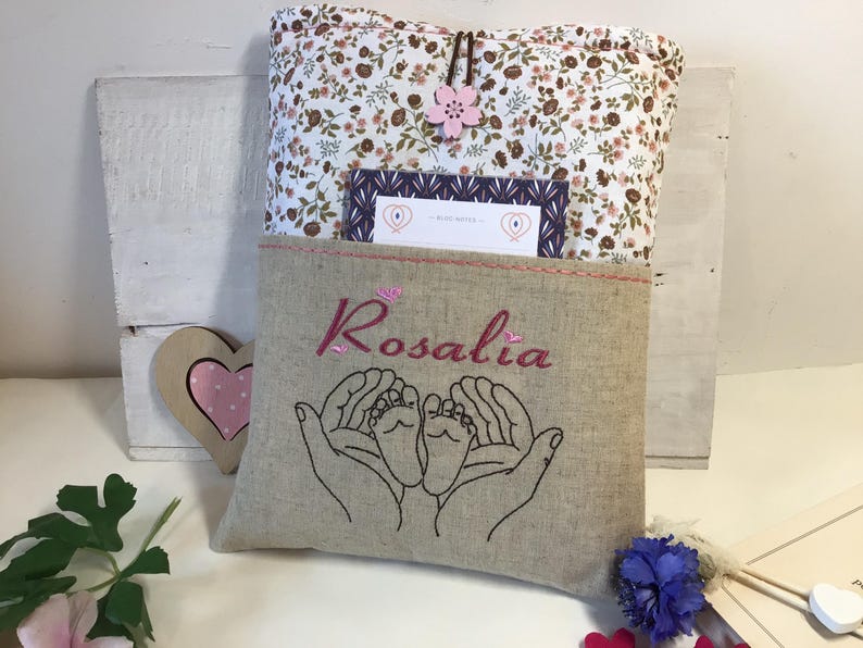 May include: A beige linen book cover with a floral pattern and a pink flower. The cover has a pocket with the name "Rosalia" embroidered in pink thread and an embroidered design of two hands holding a pair of baby feet.