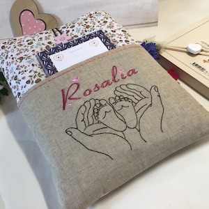 May include: A beige linen baby gift bag with a pocket. The bag is embroidered with the name "Rosalia" in pink thread and a design of two hands holding a baby's feet.