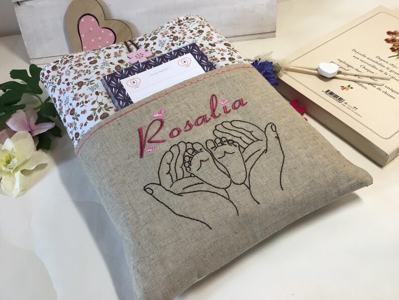 May include: A beige linen baby gift bag with a pocket. The bag is embroidered with the name "Rosalia" in pink thread and a design of two hands holding a baby's feet.