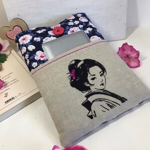K&ouml;nnte beinhalten: Eine Laptop-H&uuml;lle aus Leinen mit einem Blumendruck auf der einen Seite und einem gestickten Schwarzwei&szlig;bild einer Frau auf der anderen Seite. Die H&uuml;lle hat einen rosa Knopf und einen rosa Rei&szlig;verschluss.