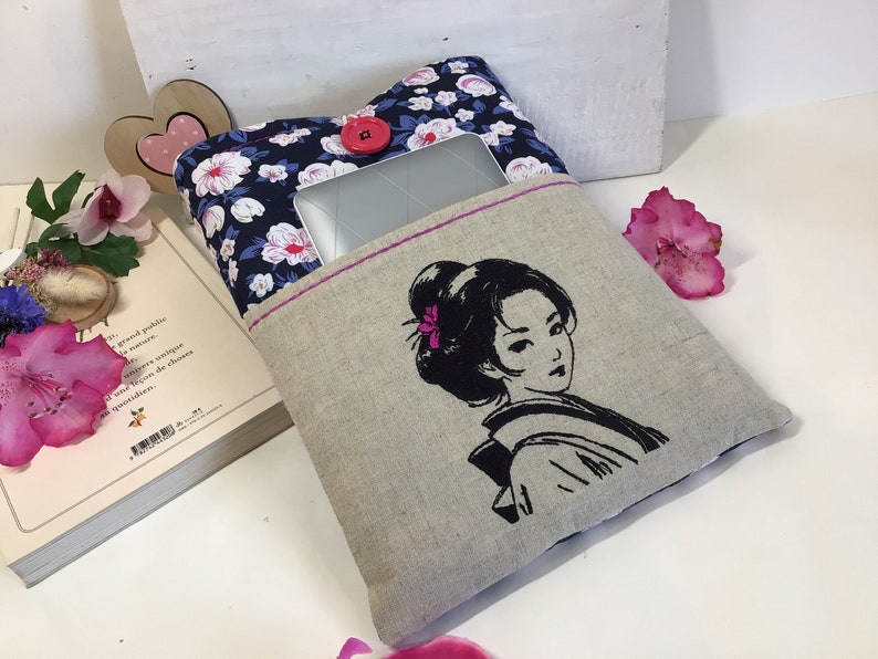 K&ouml;nnte beinhalten: Eine Laptop-H&uuml;lle aus Leinen mit einem Blumendruck auf der einen Seite und einem gestickten Schwarzwei&szlig;bild einer Frau auf der anderen Seite. Die H&uuml;lle hat einen rosa Knopf und einen rosa Rei&szlig;verschluss.
