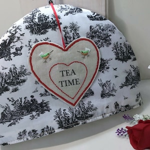 Könnte beinhalten: Ein weißer Teewärmer mit einem schwarzen Toile-de-Jouy-Muster und einer herzförmigen Applikation. Die Applikation trägt die Aufschrift "TEA TIME" und zeigt zwei gestickte Kolibris. Ein rotes Band ist zum Aufhängen befestigt.