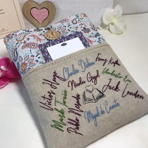 May include: A beige book sleeve with embroidered author names and a book illustration. The top features a paisley pattern and a small pocket. A wooden heart and a book are in the background.