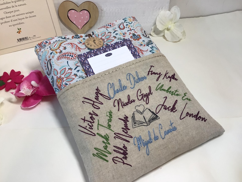 May include: A beige book sleeve with embroidered author names and a book illustration. The top features a paisley pattern and a small pocket. A wooden heart and a book are in the background.