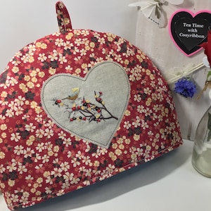 May include: Red tea cozy with a heart-shaped embroidered panel featuring a bird on a branch. The cozy has a floral pattern and a loop handle.