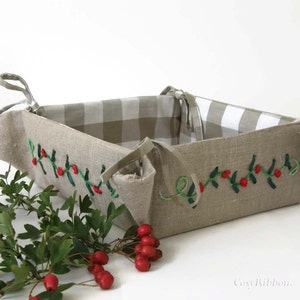 May include: A beige linen bread basket with a gingham lining and embroidered holly sprigs. The basket has two fabric ties on each side.