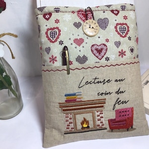 May include: A beige fabric book sleeve with a pocket, featuring embroidered text and a fireplace scene. The top section has a pattern of hearts and flowers. A pen is in the pocket. A wooden clock button secures the flap.