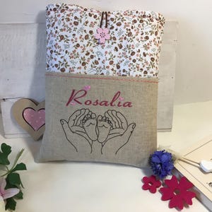 May include: A beige linen pouch with a floral print top flap. The pouch has a pink embroidered heart with two baby feet inside and the name "Rosalia" embroidered in pink.