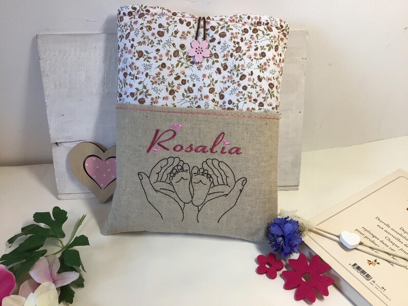 May include: A beige linen pouch with a floral print top flap. The pouch has a pink embroidered heart with two baby feet inside and the name "Rosalia" embroidered in pink.