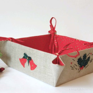 May include: A square-shaped fabric basket with red and gold fabric lining. The basket has a linen exterior with embroidered Christmas bells and holly designs. The basket has red ribbon handles.