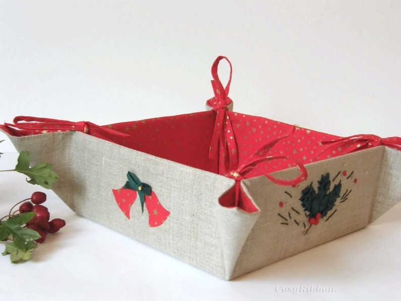 May include: A square-shaped fabric basket with red and gold fabric lining. The basket has a linen exterior with embroidered Christmas bells and holly designs. The basket has red ribbon handles.