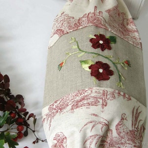 Puede incluir: Una bolsa de tela de lino beige con un patr&oacute;n de toile rojo y blanco. La bolsa tiene una cinta roja bordada con dos flores rojas y hojas verdes.