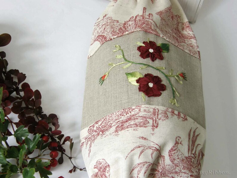Puede incluir: Una bolsa de tela de lino beige con un patr&oacute;n de toile rojo y blanco. La bolsa tiene una cinta roja bordada con dos flores rojas y hojas verdes.