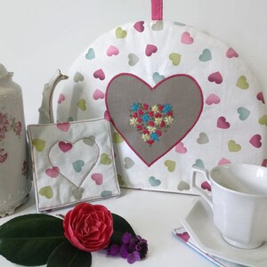 Peut inclure: Un chauffe-thé en tissu blanc avec un grand cœur brun brodé de fleurs colorées. Le chauffe-thé est décoré d'un motif de cœurs roses, verts et bleus. Il y a deux dessous-de-plat carrés assortis avec un cœur découpé.