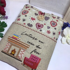 May include: Beige fabric book sleeve with embroidered fireplace, books, and armchair. The top features a pattern of hearts and snowflakes. A wooden clock button secures the flap. Text reads "Lecture au coin du feu."