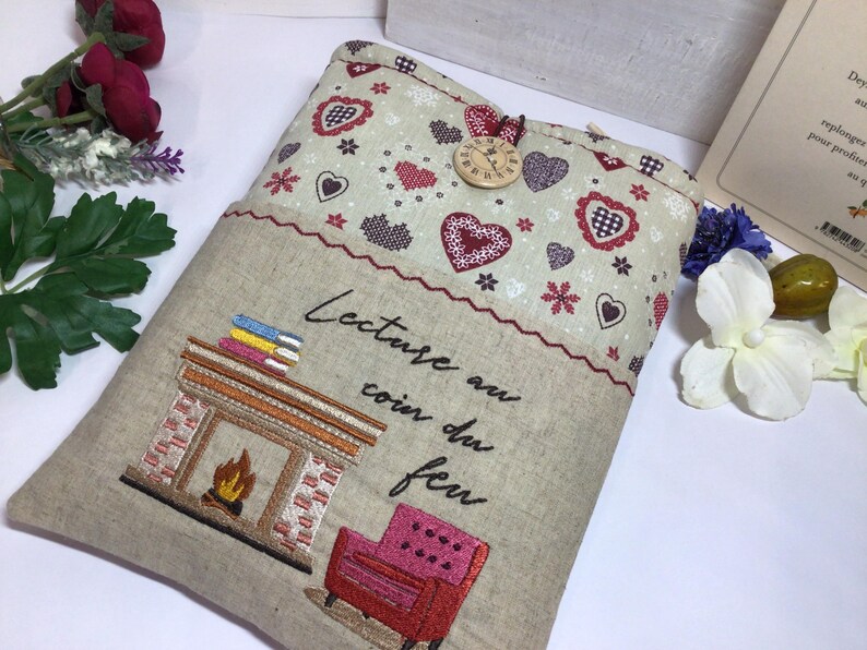 May include: Beige fabric book sleeve with embroidered fireplace, books, and armchair. The top features a pattern of hearts and snowflakes. A wooden clock button secures the flap. Text reads "Lecture au coin du feu."