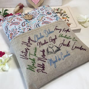 May include: A beige linen book sleeve with embroidered names of famous authors in various colors. The interior features a paisley pattern in blue, red, and white. A wooden button secures the flap.