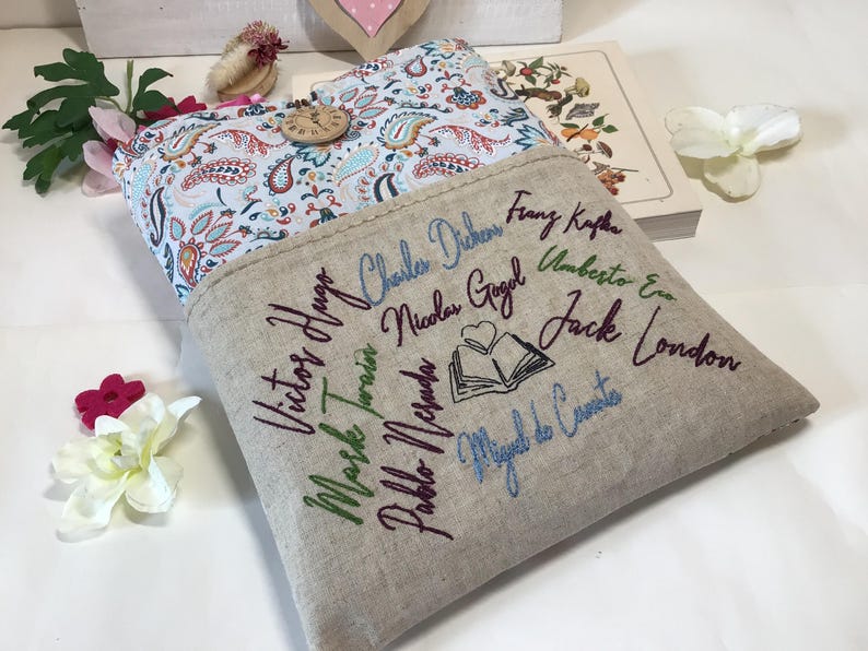 May include: A beige linen book sleeve with embroidered names of famous authors in various colors. The interior features a paisley pattern in blue, red, and white. A wooden button secures the flap.