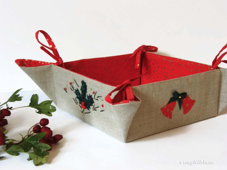 May include: A red and beige fabric storage basket with a holly and bell design. The basket has two fabric handles and is folded into a square shape.