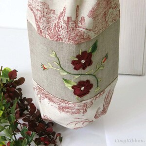 Puede incluir: Una bolsa de lino beige con un dise&ntilde;o floral rojo y blanco bordado en la parte delantera. La bolsa tiene un forro de tela con estampado rojo y blanco.