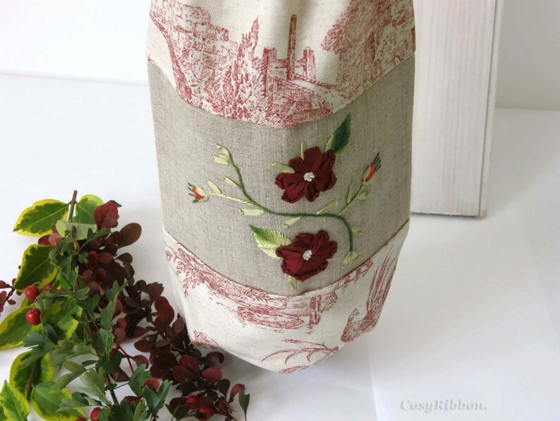 Puede incluir: Una bolsa de lino beige con un dise&ntilde;o floral rojo y blanco bordado en la parte delantera. La bolsa tiene un forro de tela con estampado rojo y blanco.