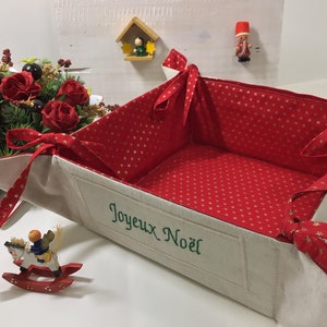 May include: A white fabric basket with red and gold star patterned lining. The basket is folded open and has a green embroidered message that says "Joyeux Noel".