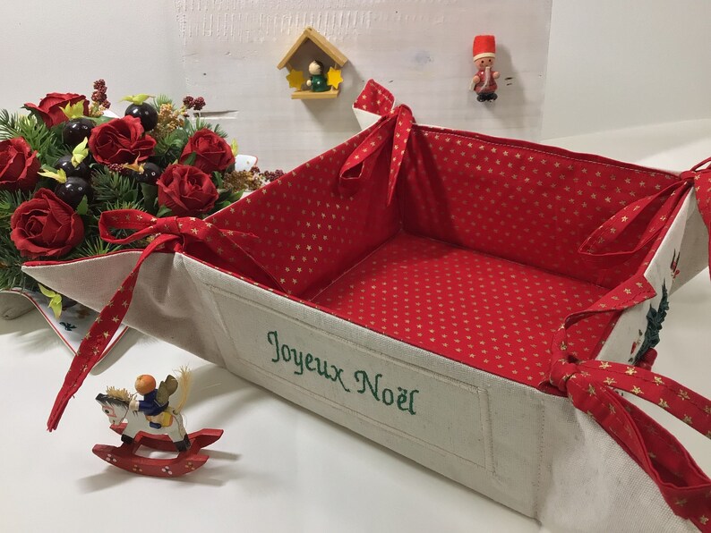 May include: A white fabric basket with red and gold star patterned lining. The basket is folded open and has a green embroidered message that says "Joyeux Noel".