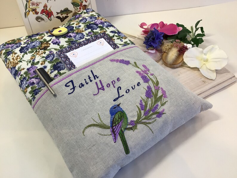 Może przedstawiać: Szara poduszka lniana z haftowanym fioletowym wzorem ptaka na gałęzi z kwiatami lawendy. Tekst "Faith Hope Love" jest wyszyty fioletowym kolorem nad ptakiem. Poduszka ma kieszeń z tyłu z długopisem w środku.