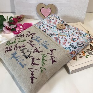 May include: Two book covers, one beige with embroidered author names and a book graphic, the other with a paisley pattern and wooden button closure. A book is partially visible. Decorative flowers and a wooden heart are in the background.