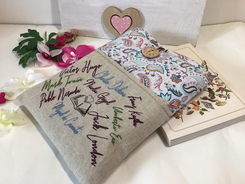 May include: Two book covers, one beige with embroidered author names and a book graphic, the other with a paisley pattern and wooden button closure. A book is partially visible. Decorative flowers and a wooden heart are in the background.