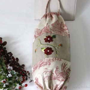 Puede incluir: Una bolsa de tela blanca y beige con cierre de cord&oacute;n. La bolsa tiene un dise&ntilde;o floral con flores rojas y hojas verdes bordadas. La bolsa est&aacute; colgada de una superficie blanca.