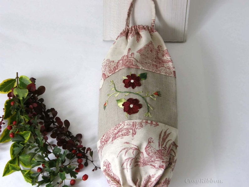 Puede incluir: Una bolsa de tela blanca y beige con cierre de cord&oacute;n. La bolsa tiene un dise&ntilde;o floral con flores rojas y hojas verdes bordadas. La bolsa est&aacute; colgada de una superficie blanca.
