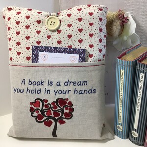 Puede incluir: Una almohada beige con un patr&oacute;n de corazones rojos y un bolsillo en la parte delantera. El bolsillo tiene una cita bordada: "Un libro es un sue&ntilde;o que se sostiene en las manos." La almohada tiene un cierre de bot&oacute;n y una peque&ntilde;a tarjeta metida en el bolsillo.