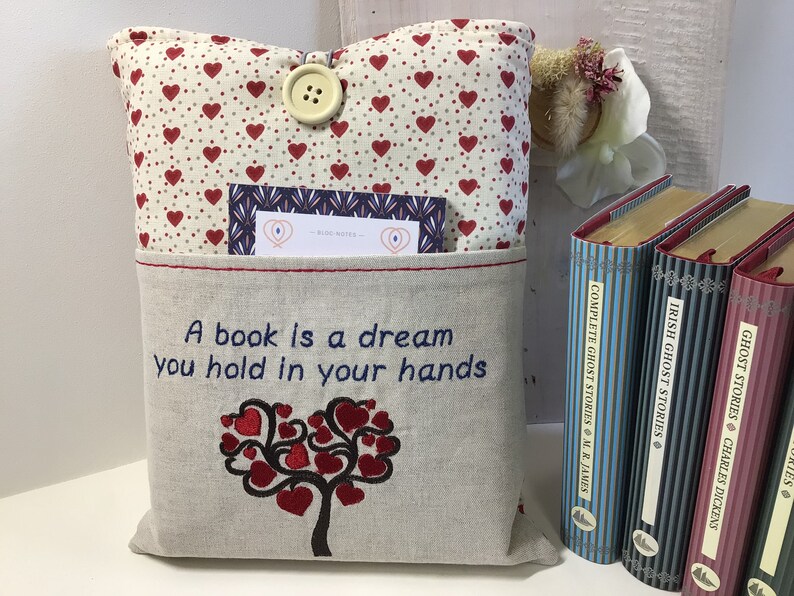 Puede incluir: Una almohada beige con un patr&oacute;n de corazones rojos y un bolsillo en la parte delantera. El bolsillo tiene una cita bordada: "Un libro es un sue&ntilde;o que se sostiene en las manos." La almohada tiene un cierre de bot&oacute;n y una peque&ntilde;a tarjeta metida en el bolsillo.