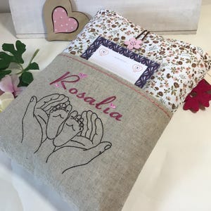 May include: A beige linen book cover with a pocket and a floral print lining. The cover is embroidered with two hands holding a pair of baby feet and the name "Rosalia".