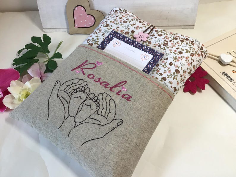 May include: A beige linen book cover with a pocket and a floral print lining. The cover is embroidered with two hands holding a pair of baby feet and the name "Rosalia".