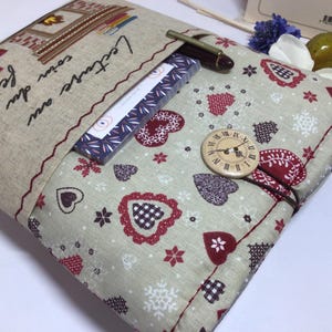 May include: A beige fabric book cover with a pocket, featuring embroidered details and the French text "Lecture au coin du feu." The cover has a pattern of red hearts and snowflakes, a wooden button with a clock face, and a pen.
