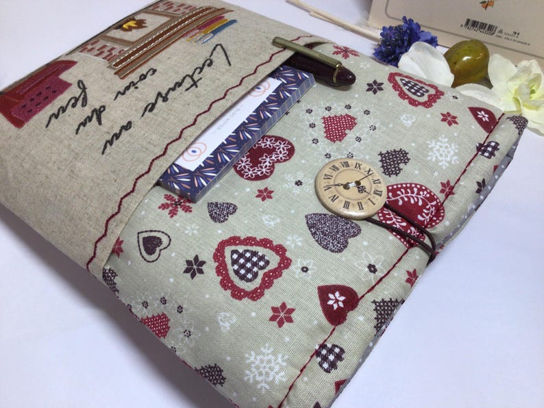 May include: A beige fabric book cover with a pocket, featuring embroidered details and the French text "Lecture au coin du feu." The cover has a pattern of red hearts and snowflakes, a wooden button with a clock face, and a pen.