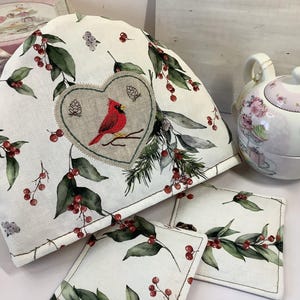 Op de afbeelding: Een witte theemuts met een hartvormige patch met een rode kardinaalvogel, dennennaalden en bessen. Een bijpassende theepot en twee onderzetters met een vergelijkbaar ontwerp zijn ook zichtbaar.