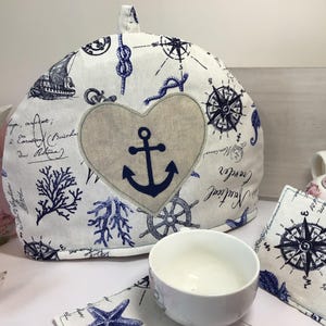 May include: A nautical-themed tea cozy with a heart-shaped anchor design. The white fabric is decorated with blue nautical illustrations, including compasses, ships, and rope knots. A white teacup and matching coasters are also visible.