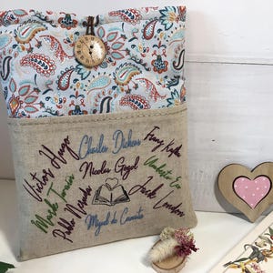 May include: A book sleeve with a paisley patterned top and a linen bottom. The bottom features embroidered names of famous authors. A wooden button and a heart-shaped decoration are also visible.