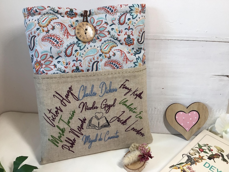 May include: A book sleeve with a paisley patterned top and a linen bottom. The bottom features embroidered names of famous authors. A wooden button and a heart-shaped decoration are also visible.