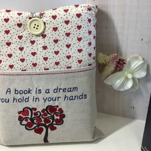 Puede incluir: Una almohada beige con un patr&oacute;n de corazones rojos en la parte delantera y un bolsillo en la parte posterior. El bolsillo tiene un dise&ntilde;o bordado de un &aacute;rbol con corazones como hojas y el texto "Un libro es un sue&ntilde;o que tienes en tus manos".
