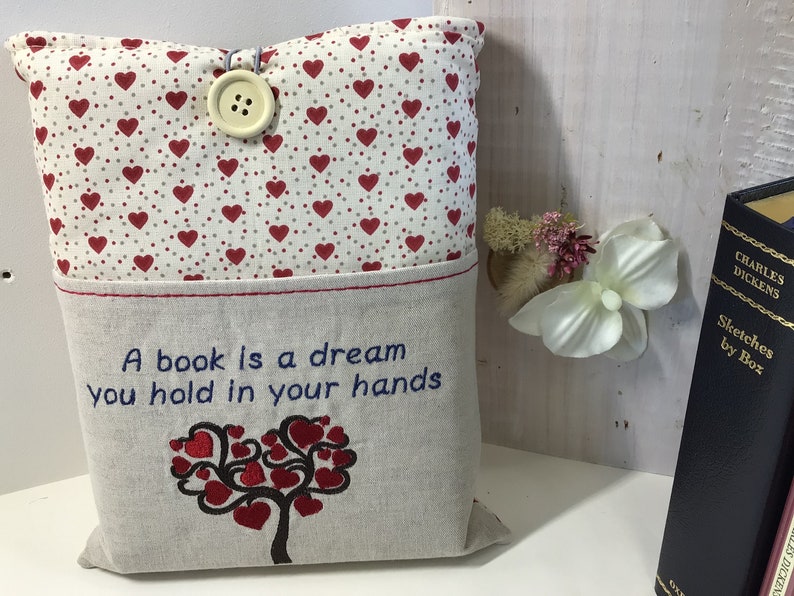 Puede incluir: Una almohada beige con un patr&oacute;n de corazones rojos en la parte delantera y un bolsillo en la parte posterior. El bolsillo tiene un dise&ntilde;o bordado de un &aacute;rbol con corazones como hojas y el texto "Un libro es un sue&ntilde;o que tienes en tus manos".