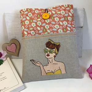 May include: A linen book cover with a pocket on the front. The pocket has an embroidered image of a woman with sunglasses and a yellow top. The top of the cover has a floral pattern with a yellow button.