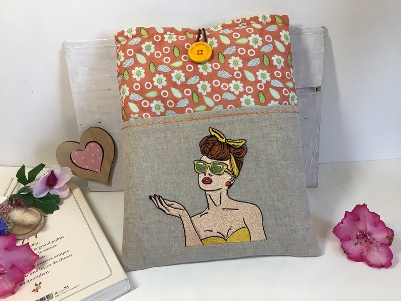 May include: A linen book cover with a pocket on the front. The pocket has an embroidered image of a woman with sunglasses and a yellow top. The top of the cover has a floral pattern with a yellow button.