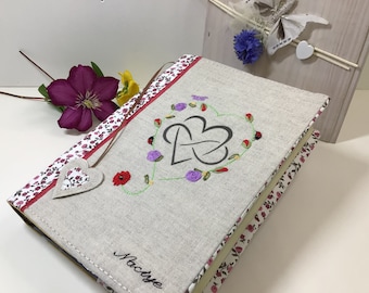 May include: A linen journal cover with a floral design and a heart with the word "Noctise" embroidered on it.