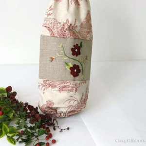 Puede incluir: Una bolsa de tela blanca con un dise&ntilde;o floral rojo y blanco. La bolsa tiene un panel de lino marr&oacute;n con flores rojas bordadas y tallos verdes. La bolsa est&aacute; hecha de una tela suave y natural y tiene un cierre de cord&oacute;n.