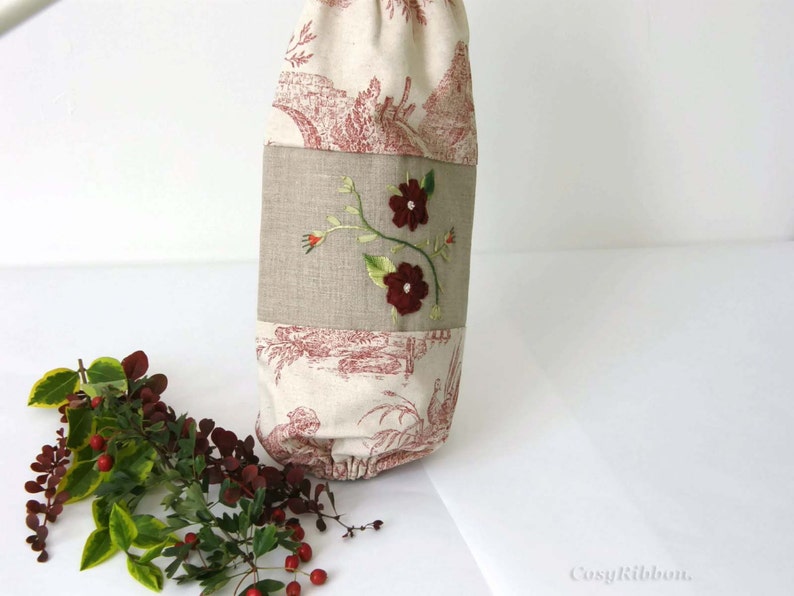 Puede incluir: Una bolsa de tela blanca con un dise&ntilde;o floral rojo y blanco. La bolsa tiene un panel de lino marr&oacute;n con flores rojas bordadas y tallos verdes. La bolsa est&aacute; hecha de una tela suave y natural y tiene un cierre de cord&oacute;n.